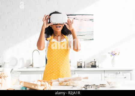 shocked african american girl in apron touching virtual reality headset at table with dough in kitchen Stock Photo