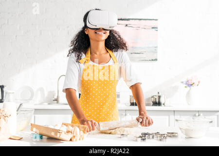 african american girl in apron and virtual reality headset rolling dough with rolling pin in kitchen Stock Photo