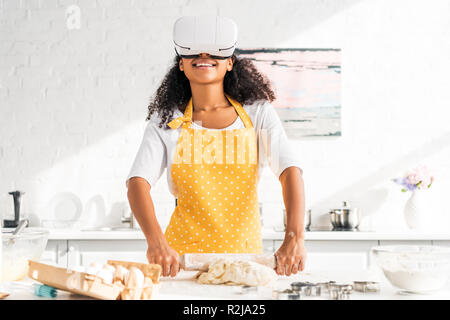 smiling african american girl in apron and virtual reality headset rolling dough in kitchen Stock Photo