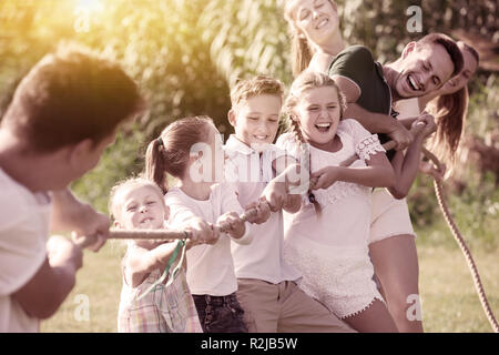 Joyful kids with moms and dads playing tug of war during joint outdoors games on sunny day Stock Photo