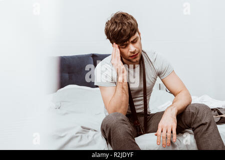 handsome young businessman with belt over neck sitting on bed and having headache at home Stock Photo
