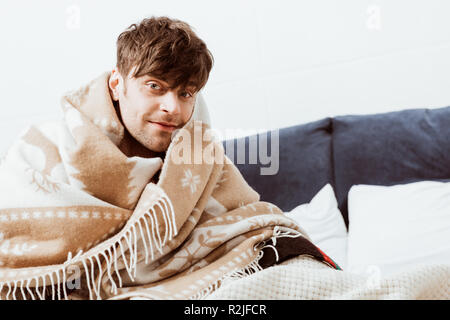 cheerful sick young man wrapped in blanket sitting on bed at home Stock Photo