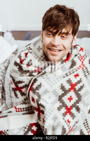 smiling sick young man wrapped in blanket sitting on bed at home Stock Photo