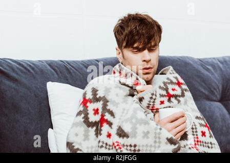 young sick man wrapped in blanket drinking tea in bed at home Stock Photo