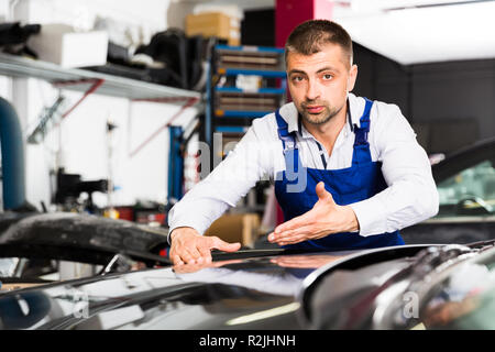 Serious male worker in blue overalls showing car after repainting at auto service Stock Photo