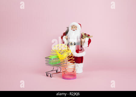 a santa claus figurine plush placed in front of a shopping trolley with rainbow spring inside on a pink background. Color harmony. Minimal still life  Stock Photo