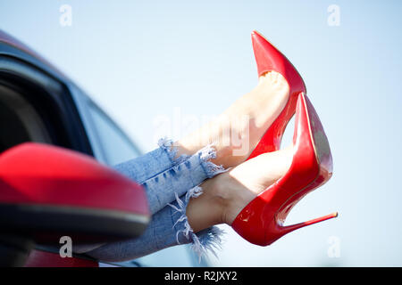 Image of girl's legs in red shoes sticking out of red car window against blue sky background Stock Photo