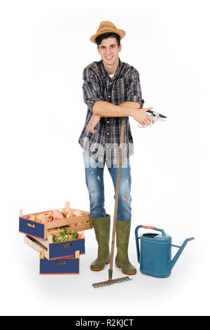Young farmer with shears in hand and rake, at his feet crates with fruit and vegetables, he is wearing a straw hat, checked shirt and green rubber boo Stock Photo