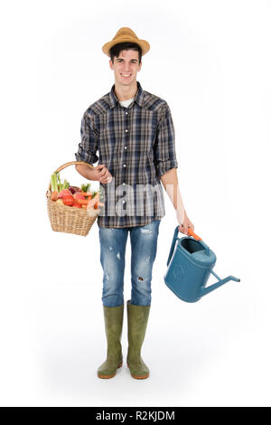 Young farmer with one arm holding a basket with fruit and vegetables, with the other a raincoat, he wears a straw hat, checked shirt and green rubber  Stock Photo