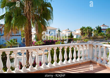 View on a street with many white villas, one high palm tree, green bushes, pink flowers and lush foliage behind a balcony and a banister. Nobody outsi Stock Photo