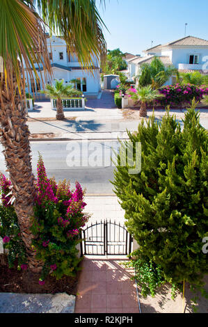 View on a street with many white villas, one high palm tree, green bushes, pink flowers and lush foliage. Entrance with a gate. Nobody outside. Warm c Stock Photo