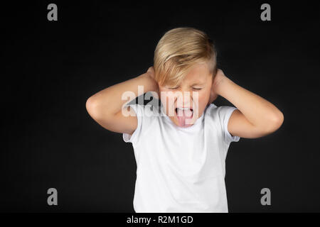 Little blond boy covers his ears and sticks out his tongue Stock Photo