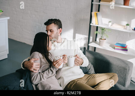 beautiful young couple with cups of cocoa with marshmallow sitting on couch at home Stock Photo
