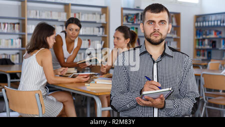 Serious young man making notes in notebook while sitting in classroom with fellow students Stock Photo