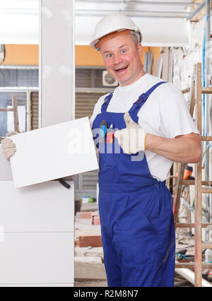 Delighted construction worker standing at indoors building site, holding white ceramic tile, giving thumbs up Stock Photo