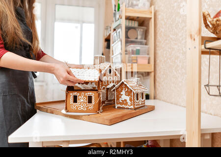 A several of biscuit gingerbread houses . Decorated with white sweet glaze. Christmas mood, winter morning. A young woman cook and packages an order or a gift Stock Photo