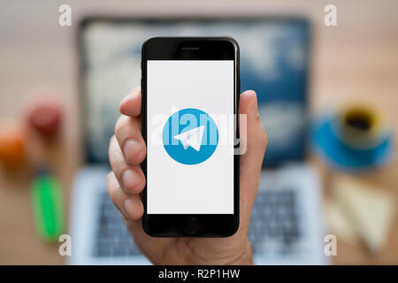 A man looks at his iPhone which displays the Telegram logo, while sat at his computer desk (Editorial use only). Stock Photo