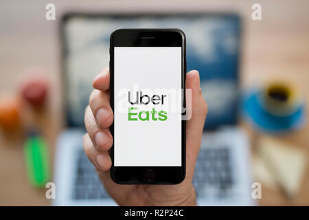 A man looks at his iPhone which displays the Uber Eats logo, while sat at his computer desk (Editorial use only). Stock Photo