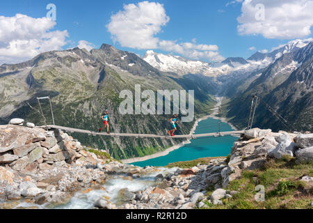 two runners on the tibetan bridge near Olperer hut with Schlegeis Stausee on the background, Zillertal Alps, Tyrol, Schwaz district, Austria. Stock Photo