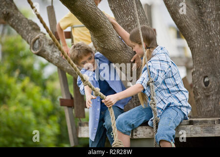 Girl handing a boy a rope swing while sitting on a wooden platform up a tree in the back yard. Stock Photo