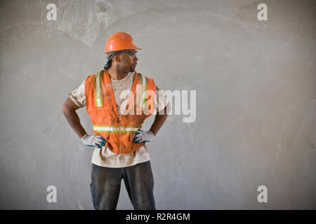 Dirty mid-adult construction worker standing with his hands on his hips while wearing a helmet and hi-visibility vest. Stock Photo