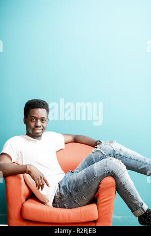 Portrait of a young african man dressed in white t-shirt and jeans sitting on the chair on the colorful background Stock Photo