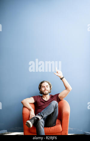 Portrait of a young caucasian bearded man with long hair showing with hands on the colorful background sitting on the chair. Image with copy space Stock Photo