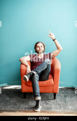 Portrait of a young caucasian bearded man with long hair showing with hands on the colorful background sitting on the chair. Image with copy space Stock Photo
