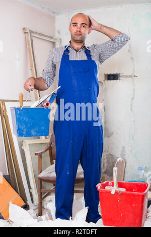 Builder is standing with bucket and putty knife and thinking about where he should start in uniform indoors. Stock Photo