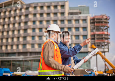 Two engineers in high visibility vests and hardhats discuss building plans on a construction site. Stock Photo