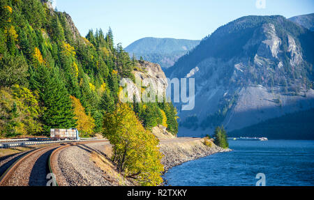 Scenic landscaping with winding railroad and highway with running semi truck with semi trailer transporting fruit boxes and river with mountain and au Stock Photo