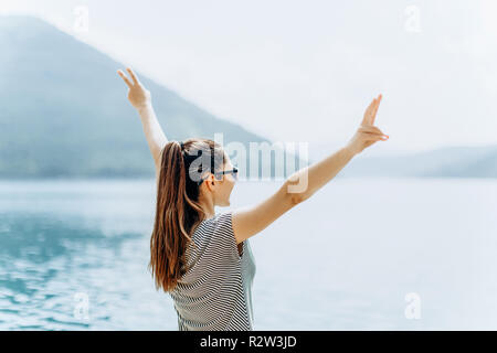 The girl on the background of the sea raises her hands up and shows how happy she is. Concept of vacation, freedom and summer time. Stock Photo