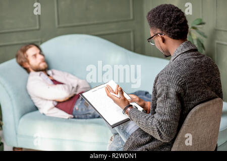 Young caucasian man visiting an african psychologist lying on the comfortable couch during psychological session in the green office Stock Photo
