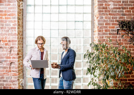 Two young businessmen dressed casually standing together with laptop near the brick wall with glass blocks indoors Stock Photo