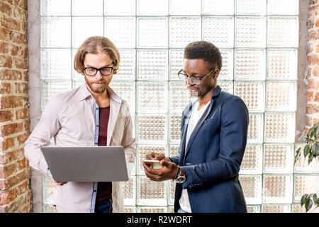 Two young businessmen dressed casually standing together with laptop near the brick wall with glass blocks indoors Stock Photo