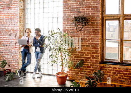 Two young businessmen dressed casually standing together near the brick wall with glass blocks in the modern office Stock Photo