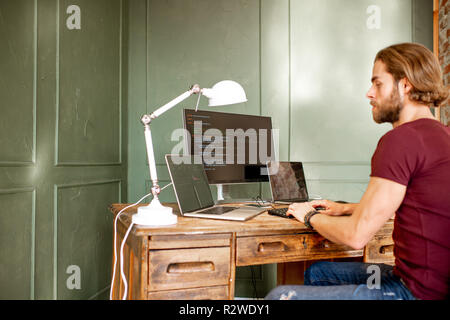 Young programmer writing a program code sitting at the workplace with three monitors in the office on the green wall background Stock Photo