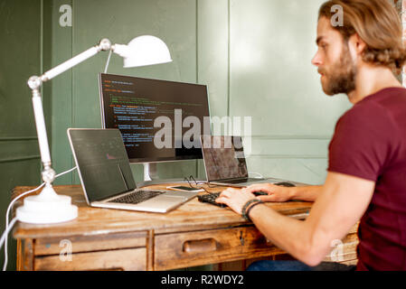 Young programmer writing a program code sitting at the workplace with three monitors in the office on the green wall background Stock Photo