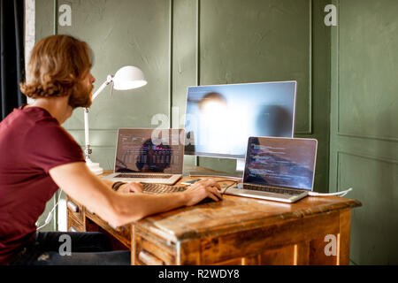 Young programmer writing a program code sitting at the workplace with three monitors in the office on the green wall background Stock Photo