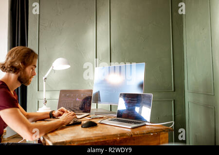 Young programmer writing a program code sitting at the workplace with three monitors in the office on the green wall background Stock Photo