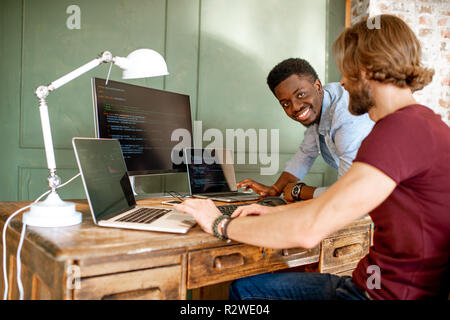 Two young programmers working with program code sitting at the workplace with three monitors in the office on the green wall background Stock Photo