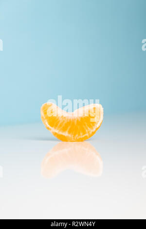 Wedge of tangerine on white and blue background. Minimalistic image of piece of orange fruit at sparse bright environment. Stock Photo