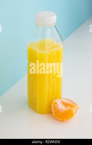 Orange drink and wedge of tangerine on white and blue background. Minimalistic image of citrus juice bottle and fruit at sparse bright environment. Stock Photo