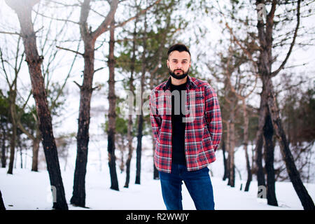 Portrait of handsome bearded young man in red plaid shirt standing in winter snowy forest. Stylish man in the woods, winter clothes collection fashion. Stock Photo