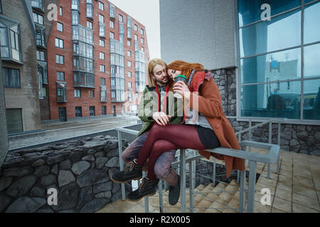 Young european couple having fun in winter urban background. Punks or hipsters style. Redhead girl is clothed in red scarf, brown coat, short shorts a Stock Photo