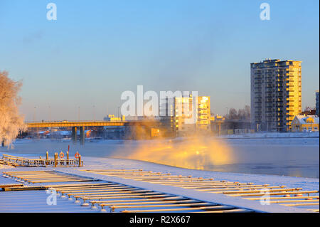 Bridge and buildings along riverbank on a cold winter day. Mist rising from freezing water. Scene lit by low angle sun. Stock Photo