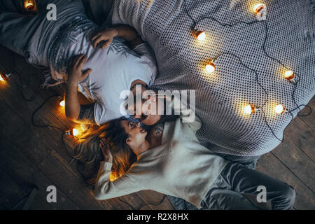 Young couple in love is lying on floor, smiling and hugging on background of glowing lightbulbs. Top view. Stock Photo