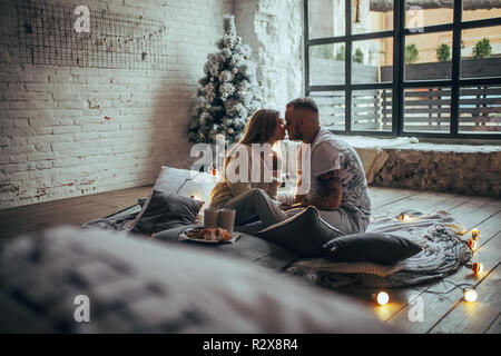 Young couple in love is sitting on floor and kissing next to two cups and plate with cookies on background of Christmas tree, glowing lightbulbs, bric Stock Photo