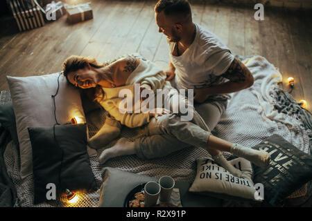 Young couple in love having fun and laughing cheerfully on background of wooden floor, coverlet, pillows and glowing lightbulbs. Stock Photo