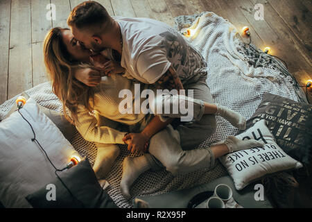 Young couple in love hugging and kissing on background of wooden floor, coverlet, pillows and glowing lightbulbs. Stock Photo